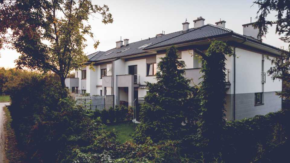 Szaro-biała elewacja budynku szeregowego Home Premium z pochyłym dachem i ogrodem, przy ul. Prostej o zmierzchu.